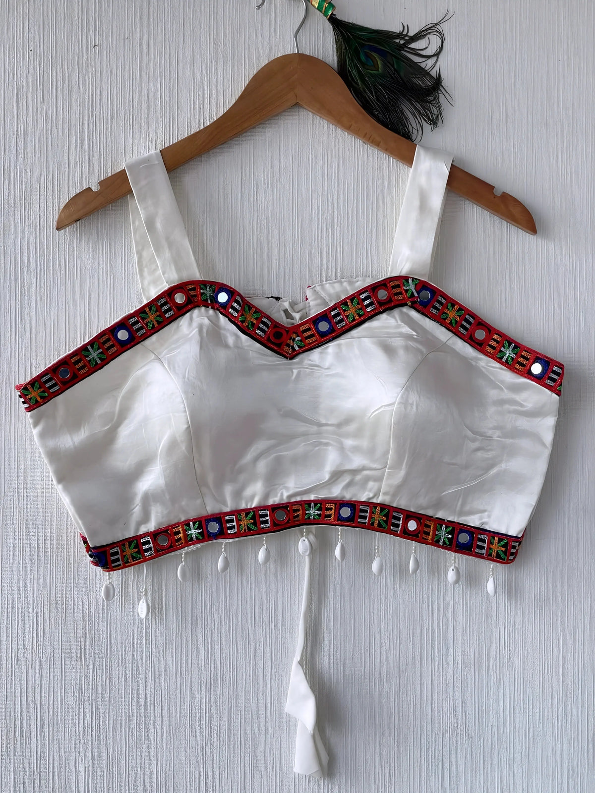 White ethnic blouse featuring vibrant mirror work and multicolored embroidered geometric border in red, green, and blue, adorned with white pearl drop tassels and a tie-up waist detail. Displayed on a wooden hanger against a textured off-white wall with peacock feathers for a festive and traditional look.