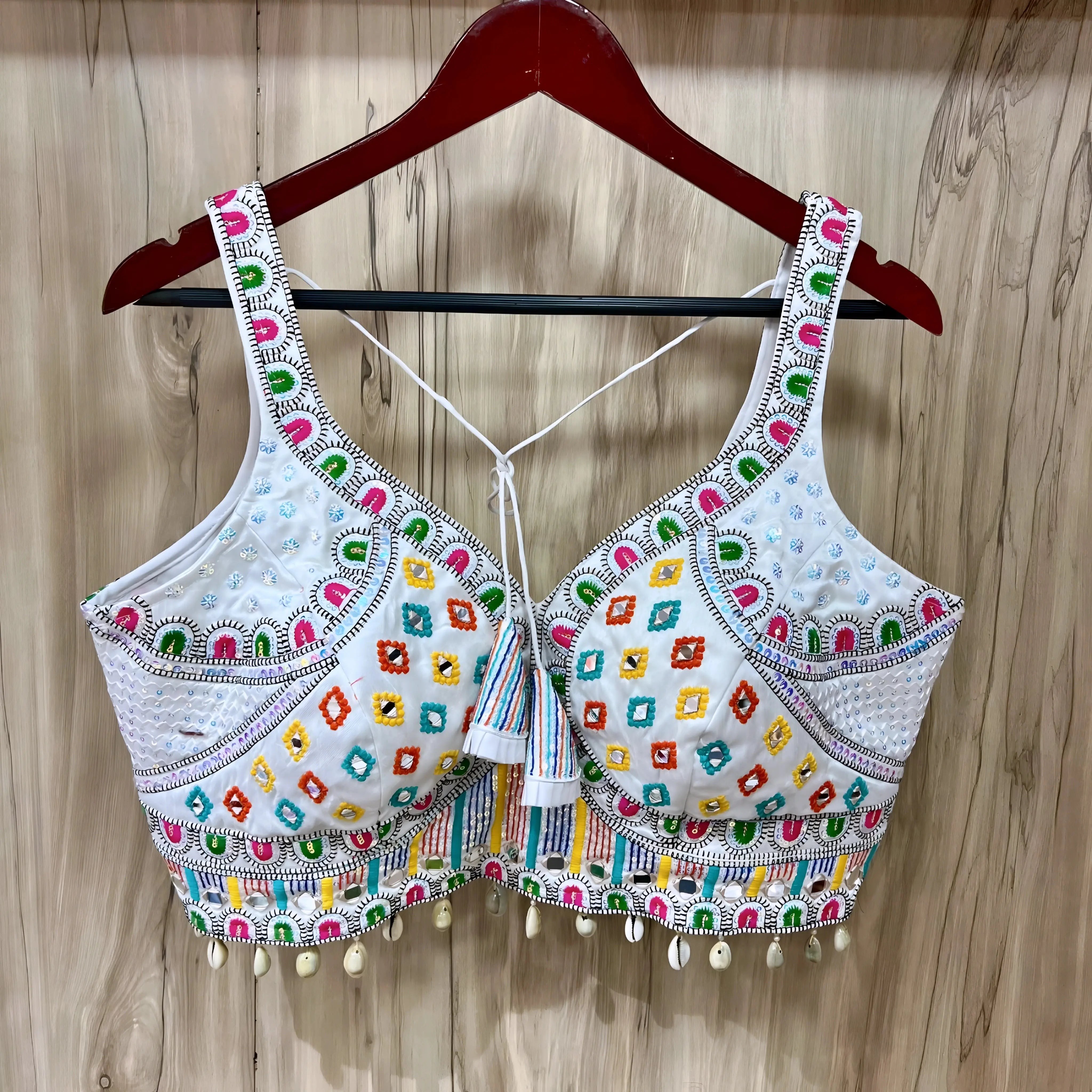 Traditional white Gujarati blouse featuring vibrant multicolor geometric embroidery in green, pink, yellow, orange, and blue, accented with intricate mirror work and white cowrie shell trim along the hem. The design includes wide embroidered straps, structured cups, tasseled tie-back detail, and is displayed on a wooden hanger against a light wood panel background.