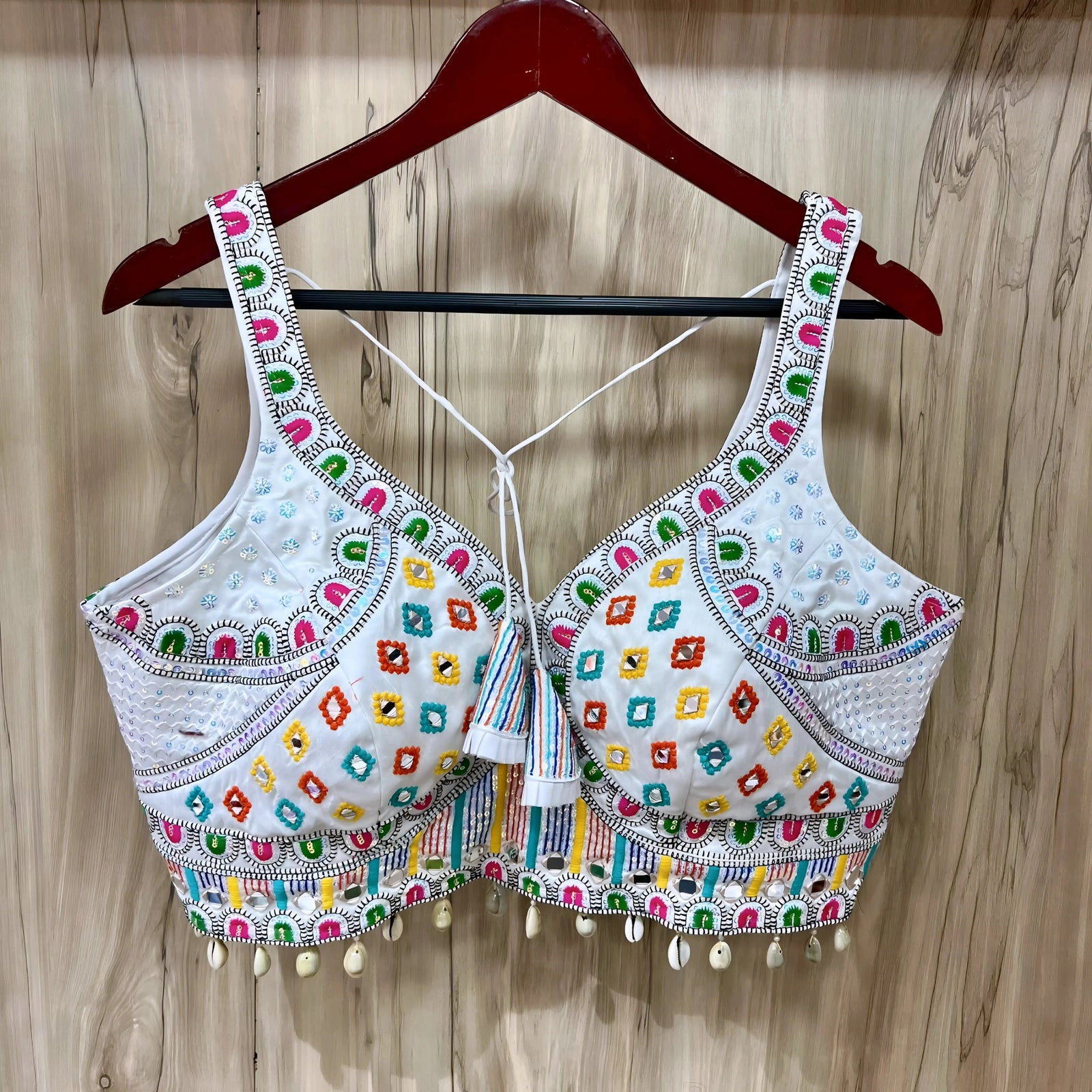 Traditional white Gujarati blouse featuring vibrant multicolor geometric embroidery in green, pink, yellow, orange, and blue, accented with intricate mirror work and white cowrie shell trim along the hem. The design includes wide embroidered straps, structured cups, tasseled tie-back detail, and is displayed on a wooden hanger against a light wood panel background.