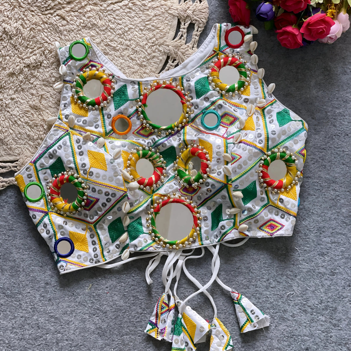 Traditional white Kutch-style blouse featuring vibrant multicolor geometric embroidery, large circular mirrors bordered with colorful beads, intricate shell accents, and tasseled tie-backs, displayed flat on a grey carpet next to a woven cream mat and a bouquet of artificial roses.