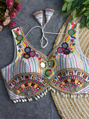 Traditional white blouse featuring vibrant multicolor mirror work, intricate thread embroidery in geometric and floral patterns, and shell tassel detailing along the hem, displayed on a flat surface with floral and leafy decor.