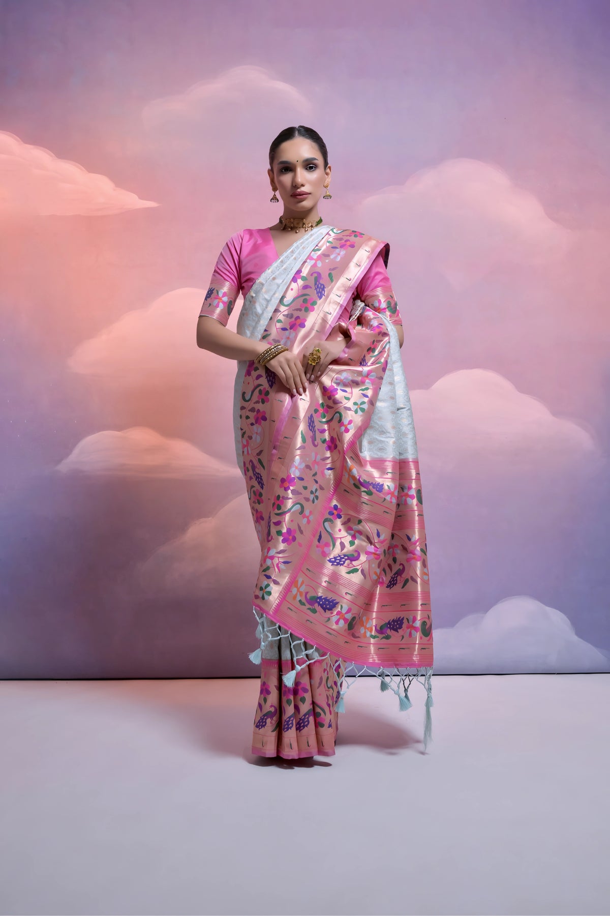 A woman models an elegant Off-White Soft Banarasi Silk Saree. The saree features a wide, vibrant Fuchsia Pink border and pallu with dense, colorful floral and vine Kanjivaram-style weaving. She wears a contrasting pink short-sleeve blouse and a gold choker necklace, posing indoors against a surreal pink and purple cloud backdrop.