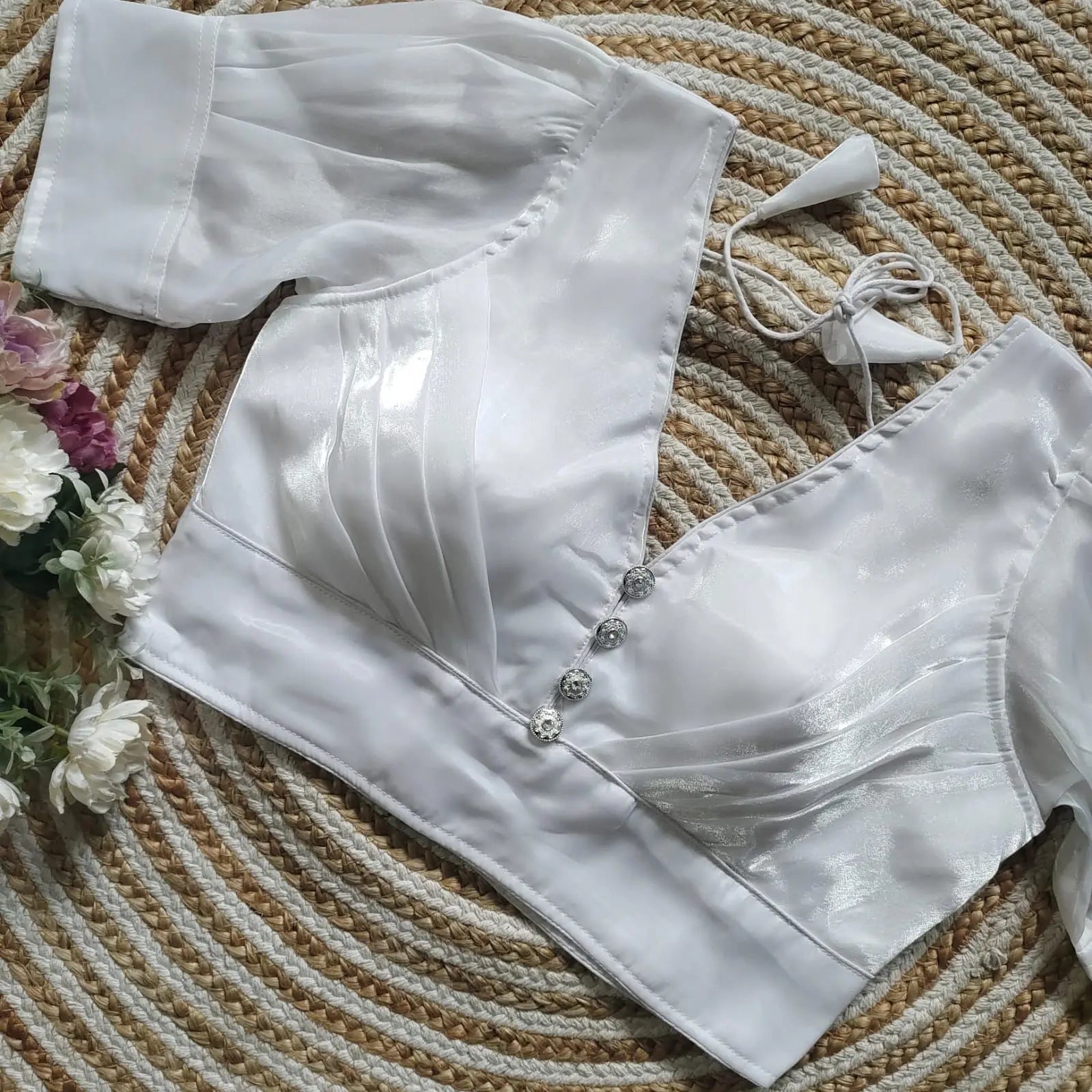 White satin blouse with pleated detailing and three decorative silver buttons on the front, featuring elbow-length sleeves and tie-up tassels, displayed on a circular woven mat with white and pink flowers on the side.