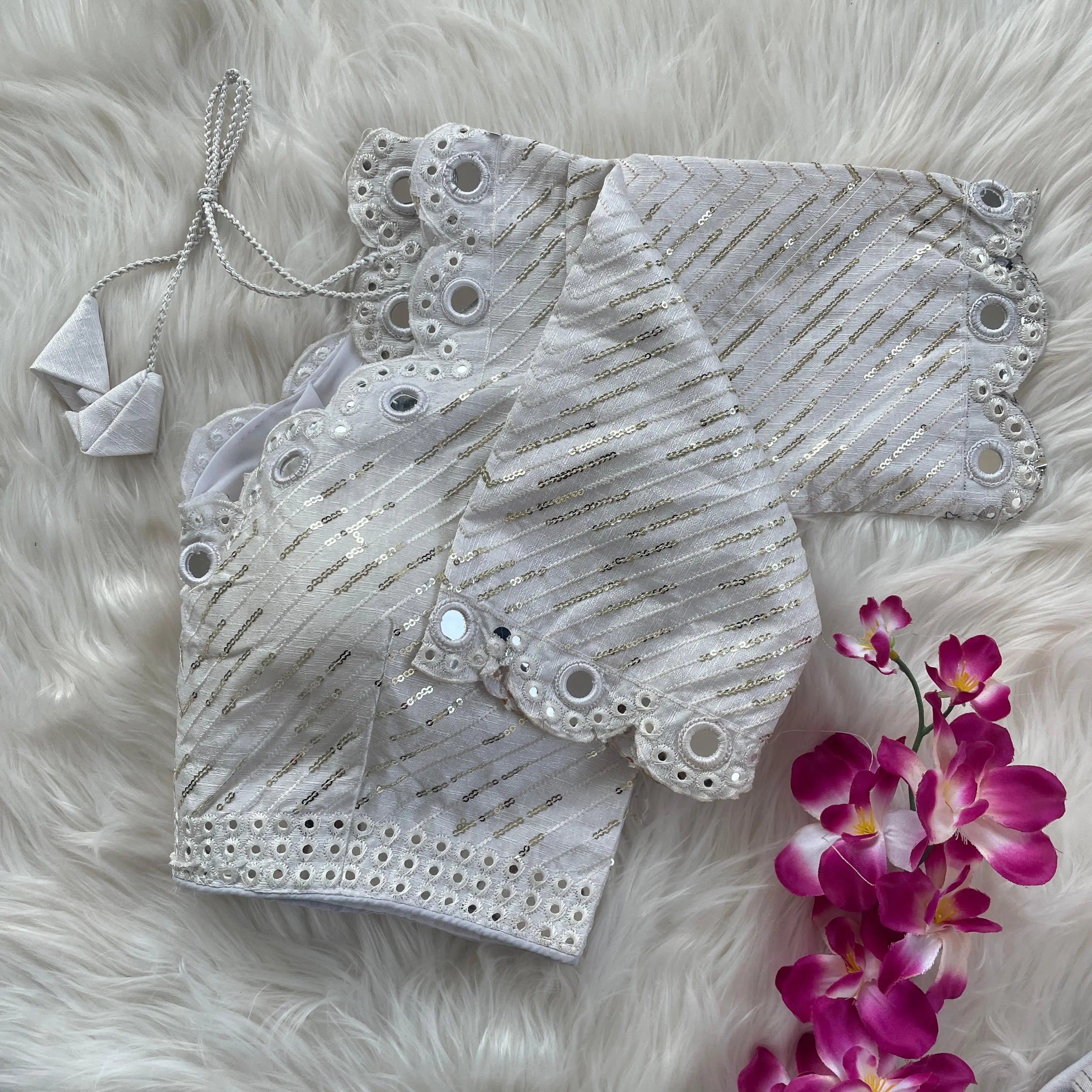 White bridal blouse with scallop sleeves, featuring circular mirror work and silver sequin stripes, decorative borders on sleeves and hem, and tie-up tassels, displayed on a textured grey and white fur background with a small potted plant nearby