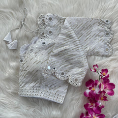 White bridal blouse with scallop sleeves, featuring circular mirror work and silver sequin stripes, decorative borders on sleeves and hem, and tie-up tassels, displayed on a textured grey and white fur background with a small potted plant nearby