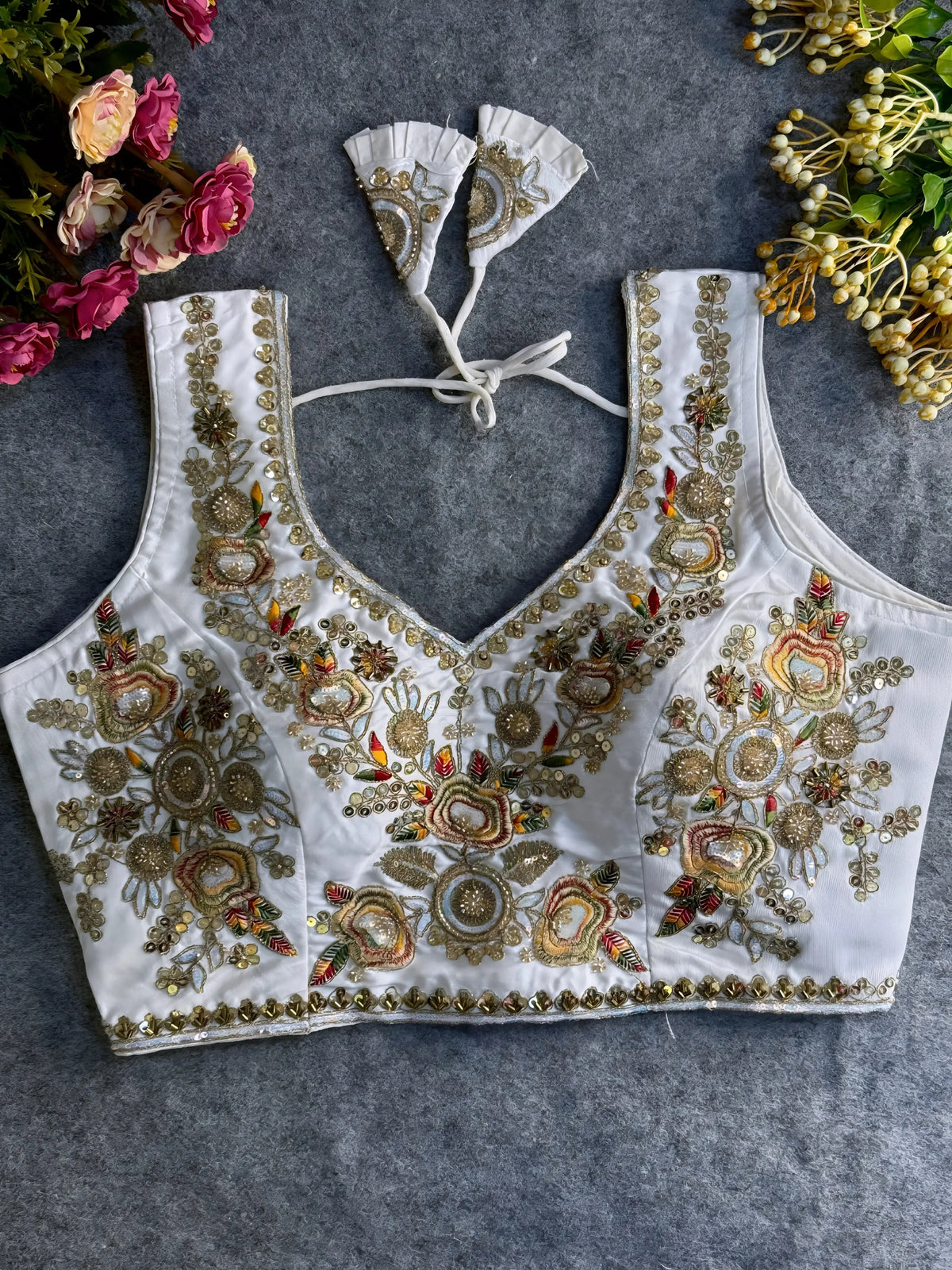  Traditional white silk blouse featuring intricate gold zardozi embroidery and beadwork with multicolor floral and leaf motifs on the front and neckline, finished with decorative tassel ties, displayed flat on a textured grey background with artificial pink and yellow flowers in the corners.