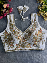  Traditional white silk blouse featuring intricate gold zardozi embroidery and beadwork with multicolor floral and leaf motifs on the front and neckline, finished with decorative tassel ties, displayed flat on a textured grey background with artificial pink and yellow flowers in the corners.