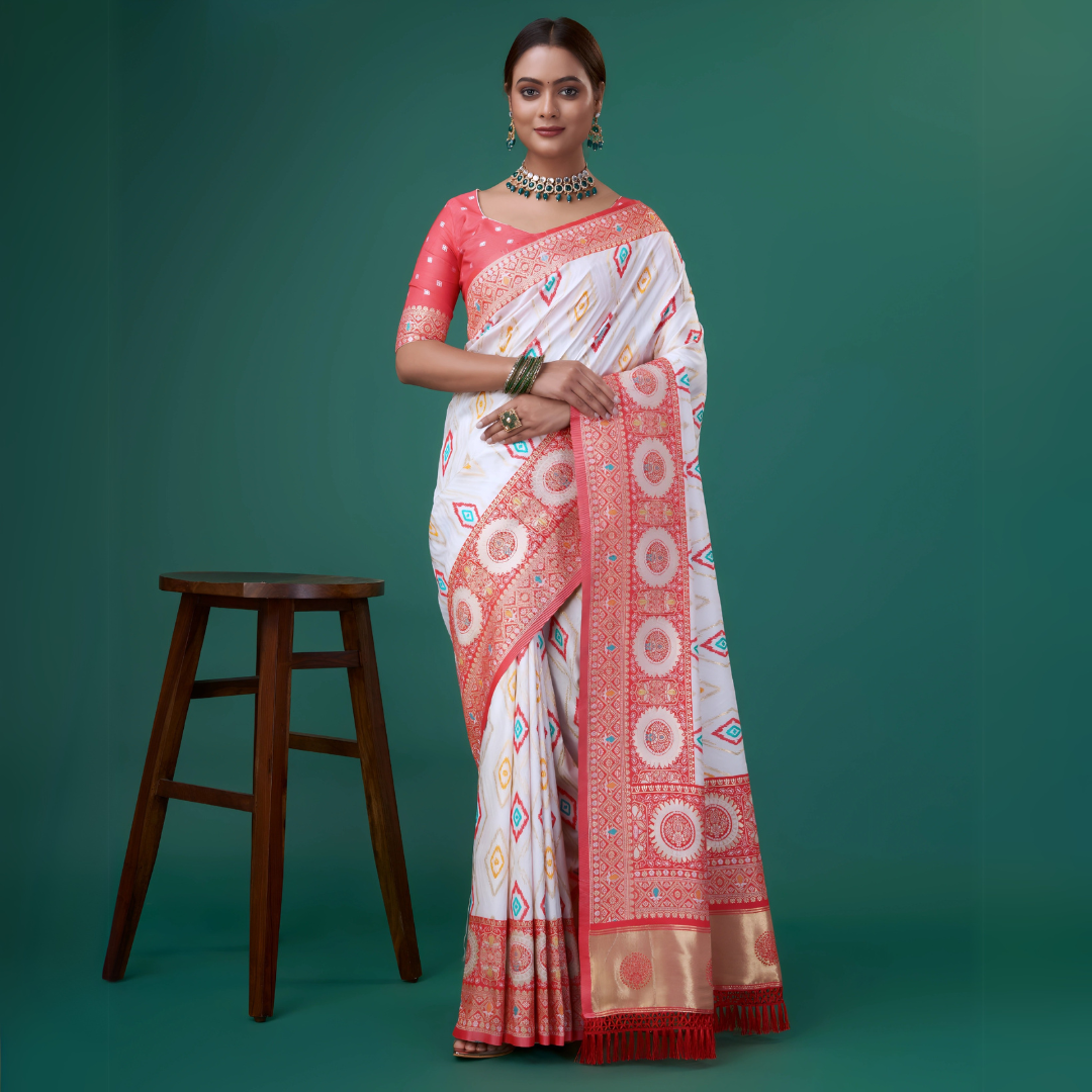 A woman models a striking White Banarasi Silk Saree featuring colorful Ikat-style motifs. The wide border and pallu are heavily woven in Coral Red Zari with intricate traditional medallion patterns. She wears a coral red blouse and a heavy green gemstone choker necklace.