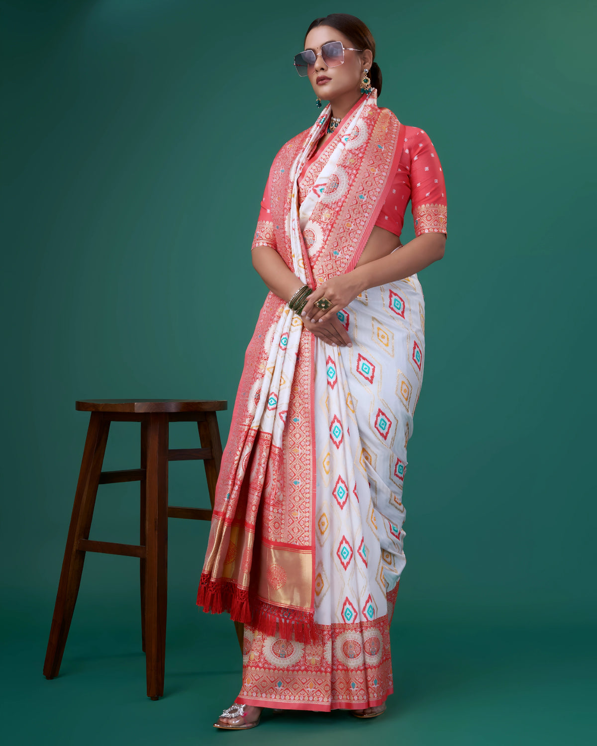 A woman models a striking White Banarasi Silk Saree featuring colorful Ikat-style motifs. The wide border and pallu are heavily woven in Coral Red Zari with intricate traditional medallion patterns. She wears a coral red blouse and a heavy green gemstone choker necklace.