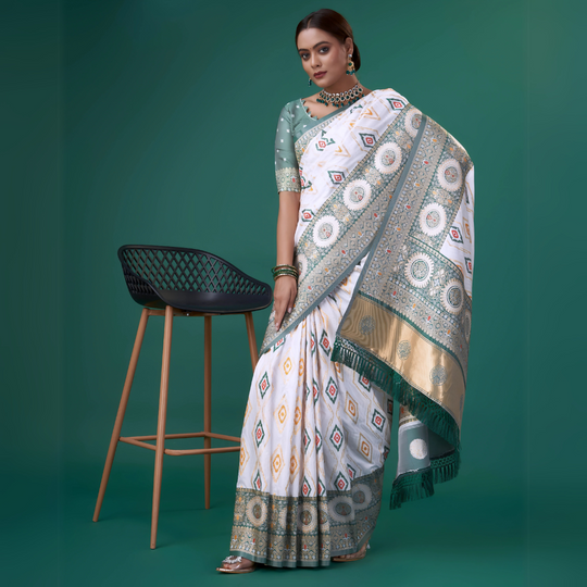 A woman models a striking White Patola Silk Saree featuring colorful Ikat-style motifs. The wide border and pallu are heavily woven in sage green Zari with intricate traditional medallion patterns. She wears a light green blouse and a heavy green gemstone choker necklace.