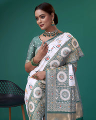 A woman models a striking White Patola Silk Saree featuring colorful Ikat-style motifs. The wide border and pallu are heavily woven in sage green Zari with intricate traditional medallion patterns. She wears a light green blouse and a heavy green gemstone choker necklace.