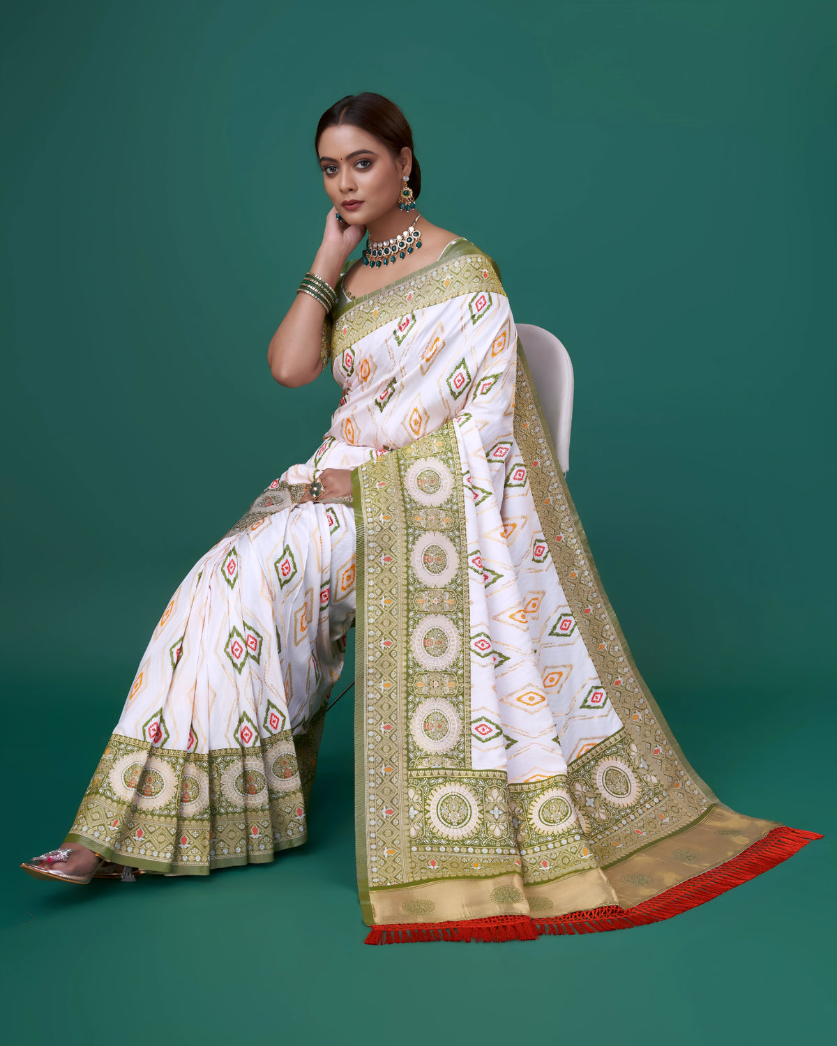 A woman models a striking White Patola Silk Saree featuring colorful Ikat-style motifs. The wide border and pallu are heavily woven in sage green Zari with intricate traditional medallion patterns. She wears a light green blouse and a heavy green gemstone choker necklace.