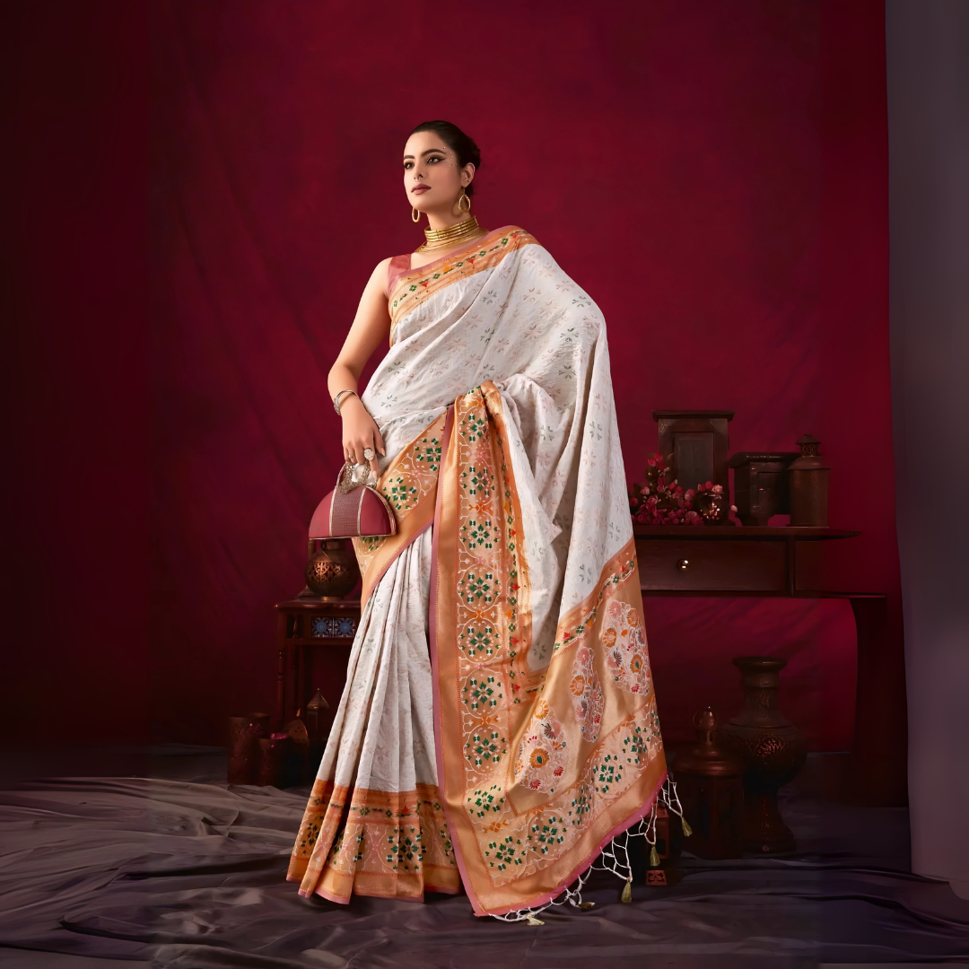 A woman models an elegant Off-White Soft Banarasi Silk Saree. The saree has a wide, prominent border and pallu with rich gold Zari weaving/print in shades of orange, peach, and green. She wears a deep maroon sleeveless blouse, gold Kundan jewelry, and carries a dome-shaped handbag, posing indoors against a dark red backdrop.
