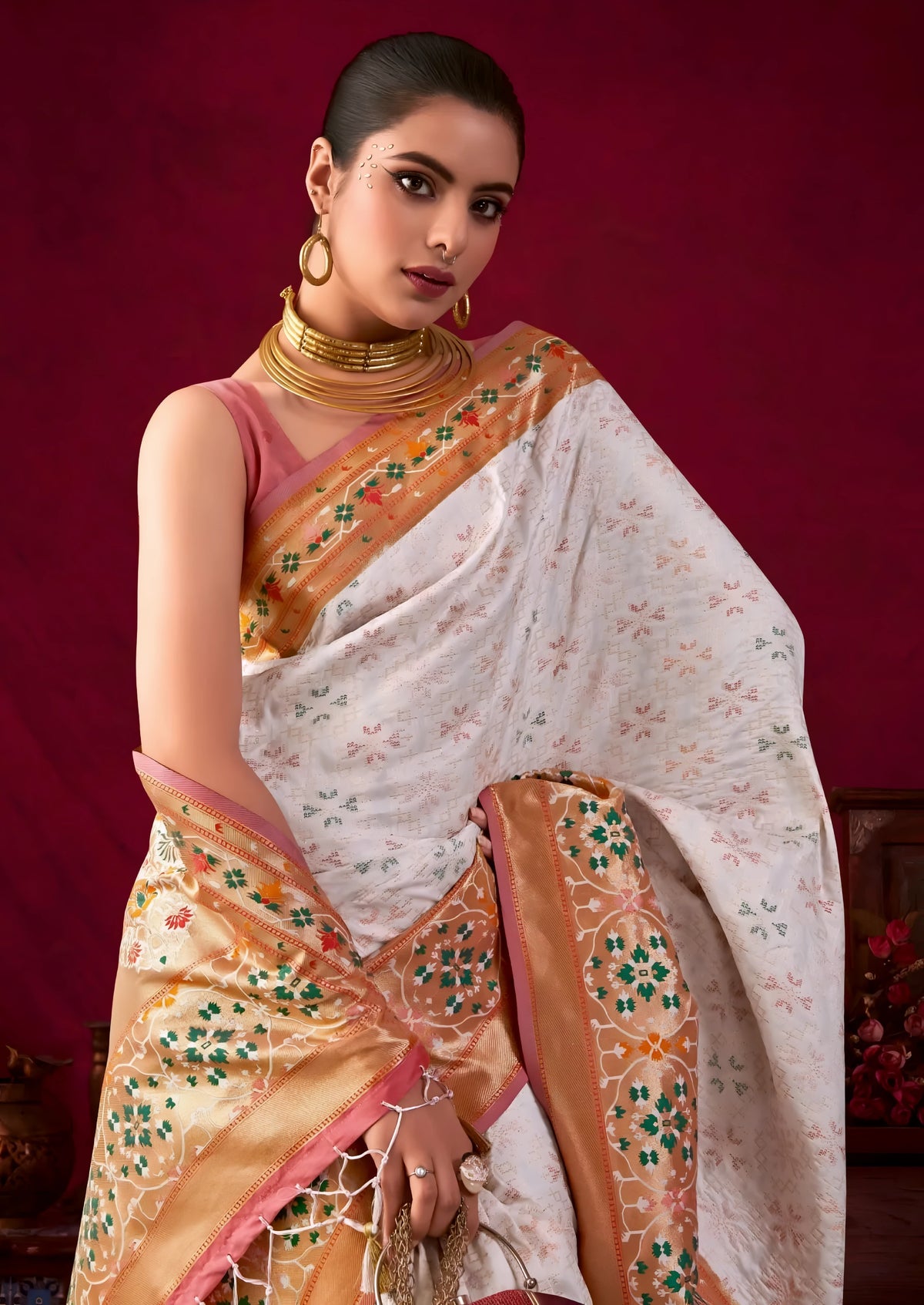 A woman models an elegant Off-White Soft Banarasi Silk Saree. The saree has a wide, prominent border and pallu with rich gold Zari weaving/print in shades of orange, peach, and green. She wears a deep maroon sleeveless blouse, gold Kundan jewelry, and carries a dome-shaped handbag, posing indoors against a dark red backdrop.