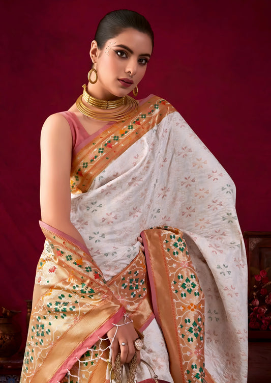 A woman models an elegant Off-White Soft Banarasi Silk Saree. The saree has a wide, prominent border and pallu with rich gold Zari weaving/print in shades of orange, peach, and green. She wears a deep maroon sleeveless blouse, gold Kundan jewelry, and carries a dome-shaped handbag, posing indoors against a dark red backdrop.