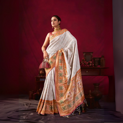 A woman models an elegant Off-White Soft Banarasi Silk Saree. The saree has a wide, prominent border and pallu with rich gold Zari weaving/print in shades of orange, peach, and green. She wears a deep maroon sleeveless blouse, gold Kundan jewelry, and carries a dome-shaped handbag, posing indoors against a dark red backdrop.