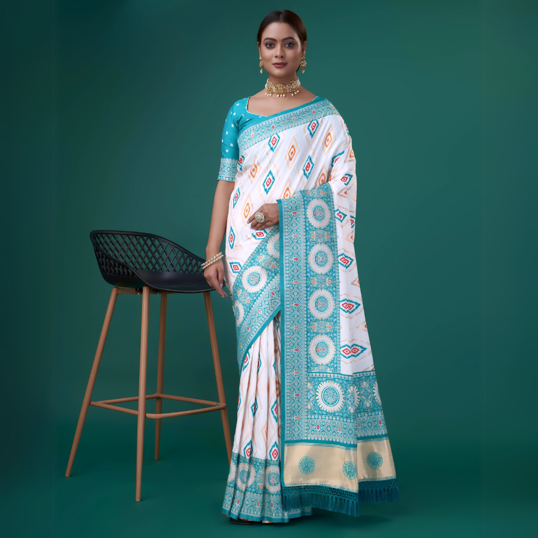 A woman models a striking White Patola Silk Saree featuring Ikat-style diamond motifs. The wide border and pallu are heavily woven in peacock blue Zari with intricate geometric patterns. She wears a blue blouse and heavy gold and pearl jewelry.