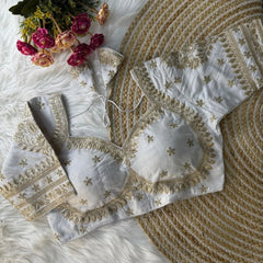 White blouse with intricate gold zari embroidery featuring floral and leaf motifs, decorative borders on sleeves and neckline, and tie-up tassels, displayed on a textured mat with pink flowers in the background