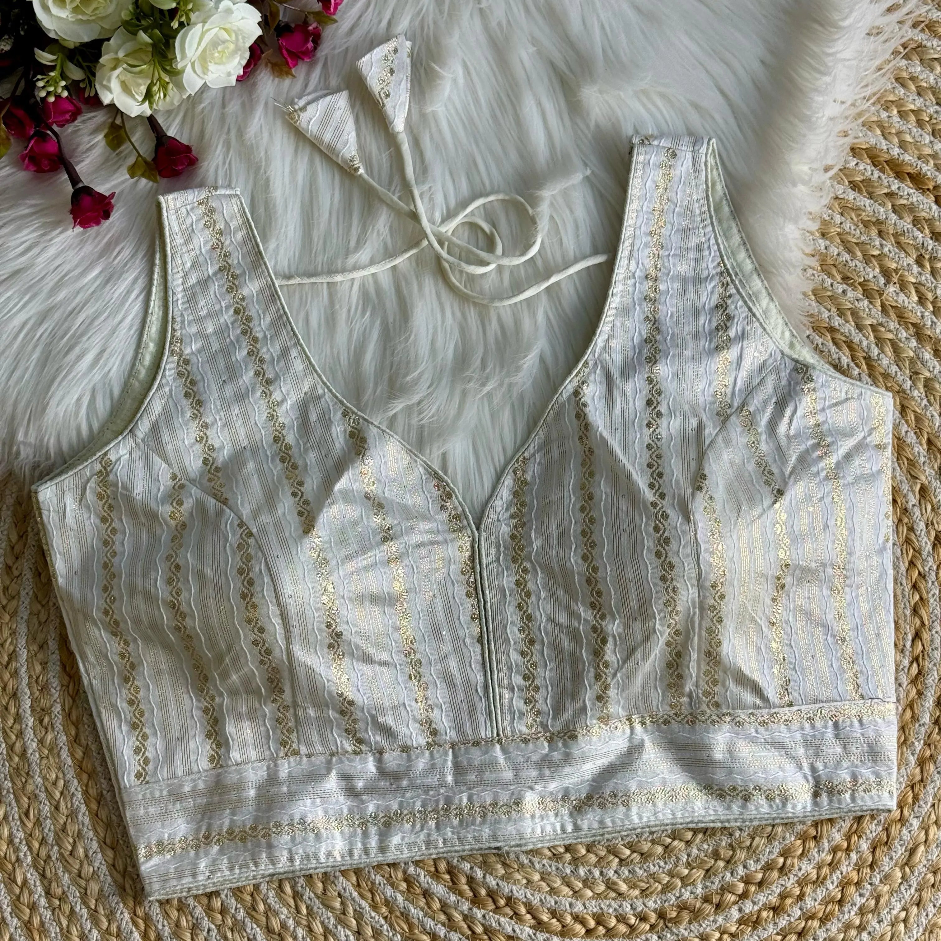 White blouse top with intricate zari work design, featuring delicate embroidery patterns, displayed on a textured background with floral accents.