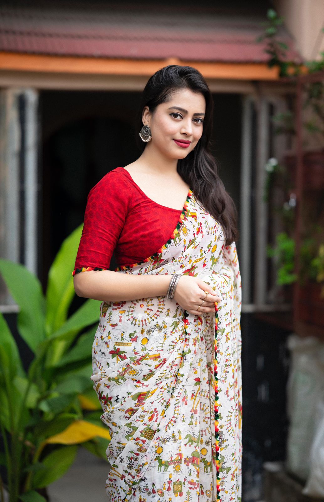Woman in a white zari work saree with pom-pom border standing on a tiled patio with lush greenery and a traditional house in the background