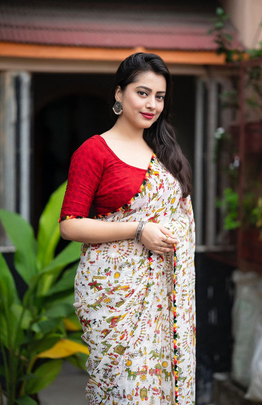 Woman in a white zari work saree with pom-pom border standing on a tiled patio with lush greenery and a traditional house in the background
