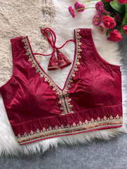  Traditional wine red silk blouse featuring intricate gold embroidery and beadwork along the neckline and hem, with a decorative tassel tie at the back, displayed flat on a soft fur and textured mat background with artificial pink flowers in the corner.
