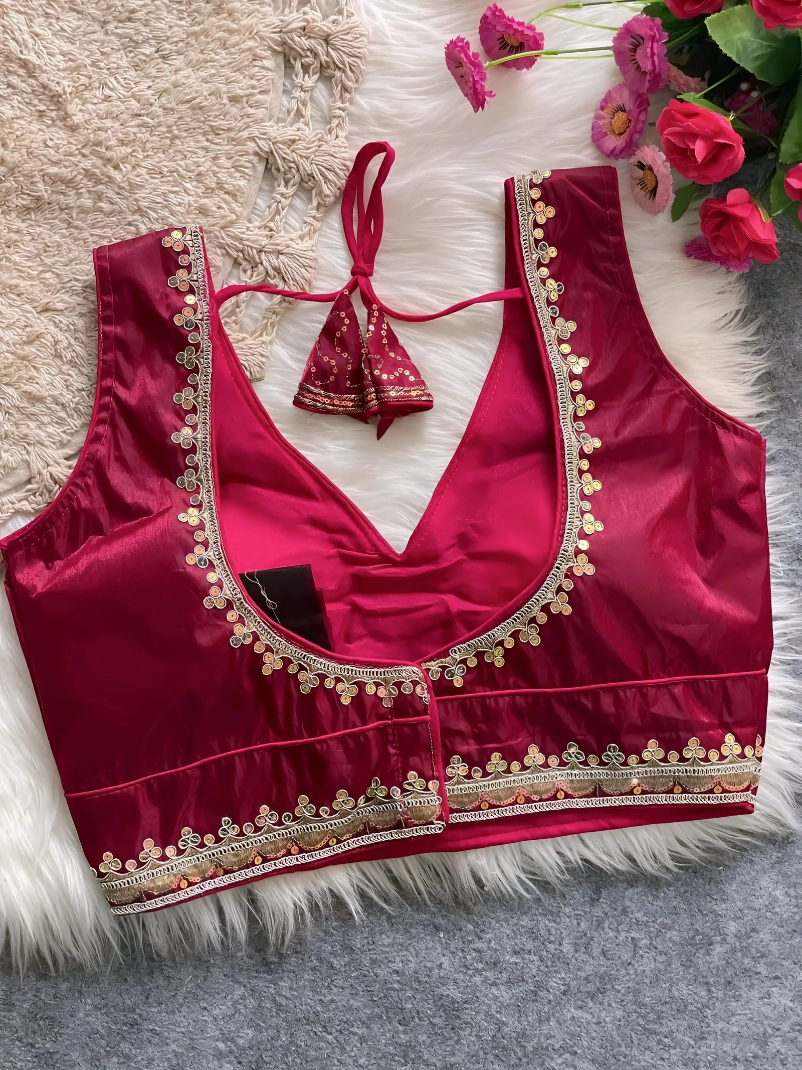  Traditional wine red silk blouse featuring intricate gold embroidery and beadwork along the neckline and hem, with a decorative tassel tie at the back, displayed flat on a soft fur and textured mat background with artificial pink flowers in the corner.