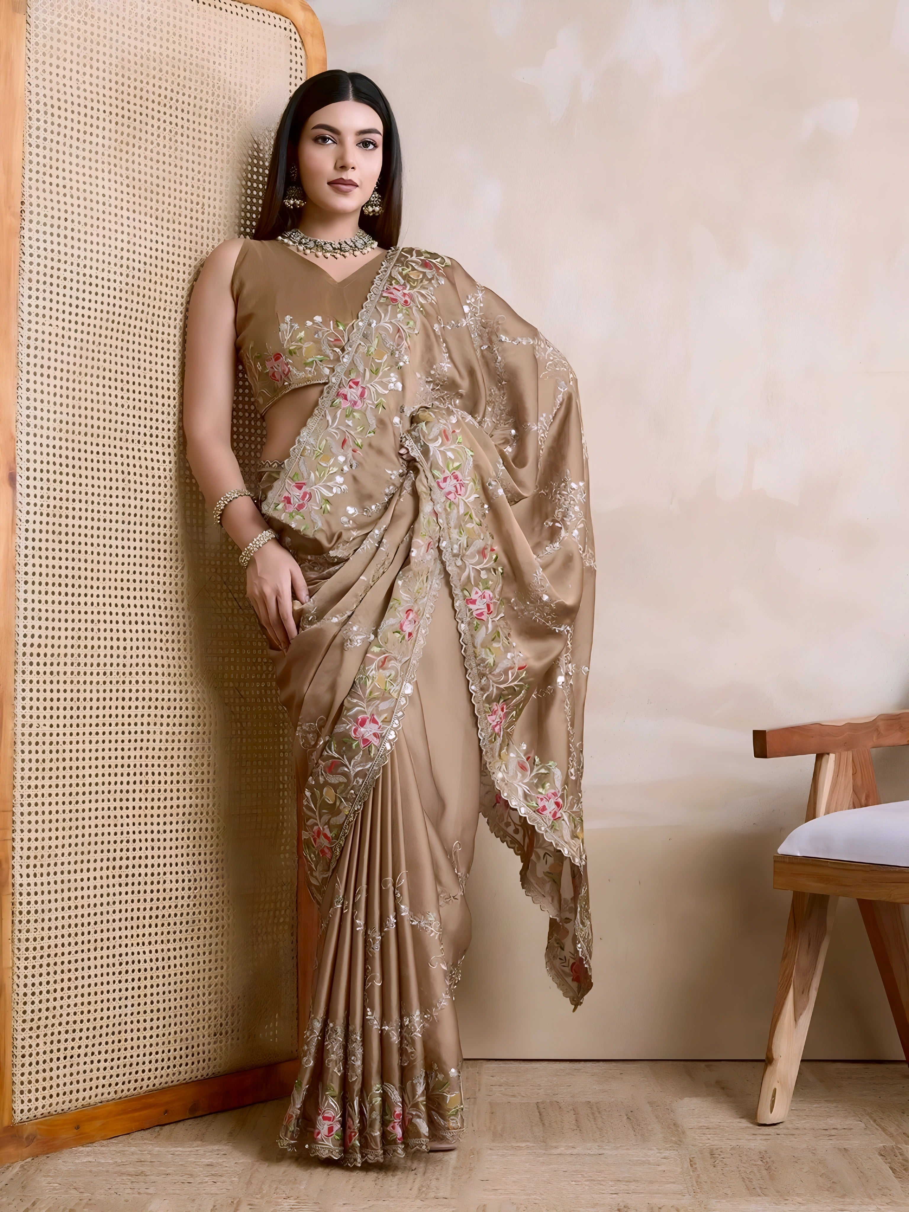 A woman models an opulent Beige Rangoli Silk Saree with scattered sequins. The saree is bordered by wide, dense floral embroidery in pink, red, and green threadwork. She wears a beige sleeveless blouse and a heavy green gemstone necklace.
