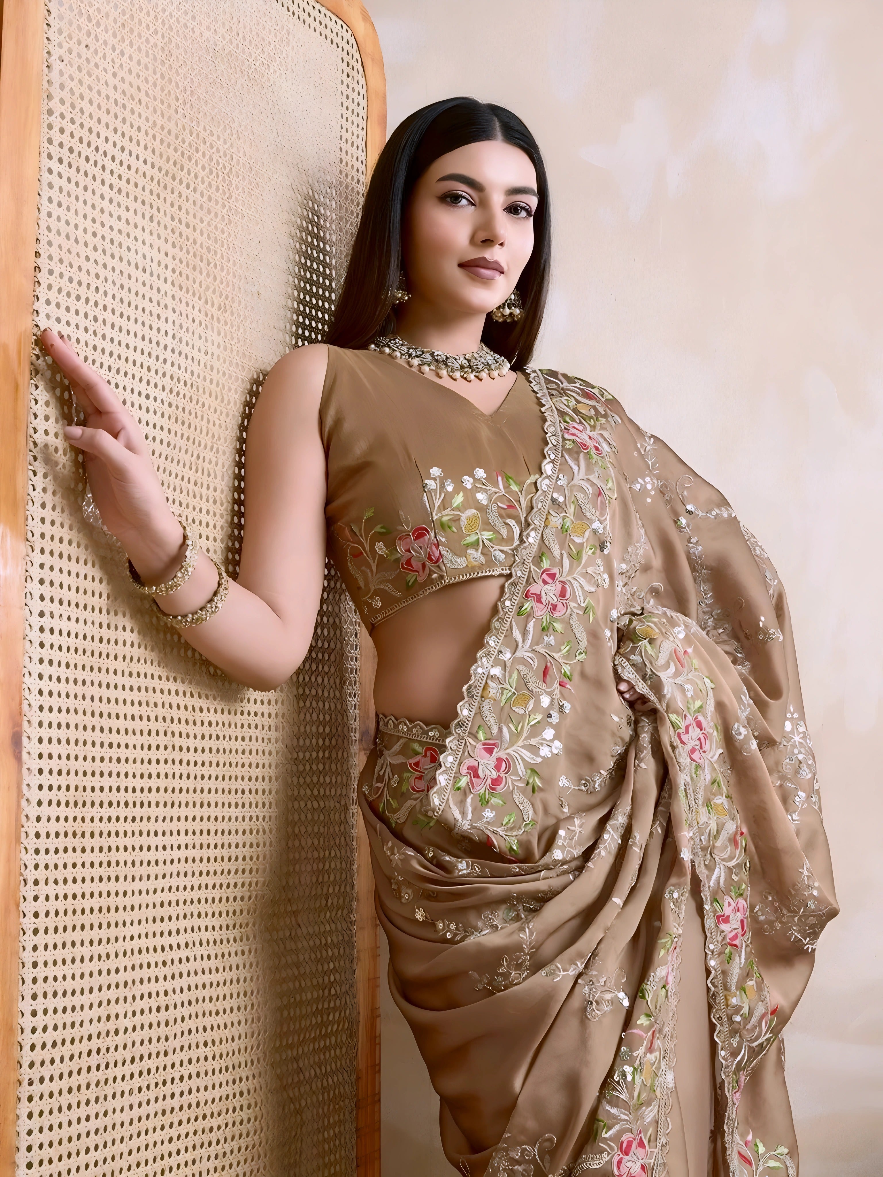 A woman models an opulent Beige Rangoli Silk Saree with scattered sequins. The saree is bordered by wide, dense floral embroidery in pink, red, and green threadwork. She wears a beige sleeveless blouse and a heavy green gemstone necklace.