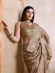 A woman models an opulent Beige Rangoli Silk Saree with scattered sequins. The saree is bordered by wide, dense floral embroidery in pink, red, and green threadwork. She wears a beige sleeveless blouse and a heavy green gemstone necklace.