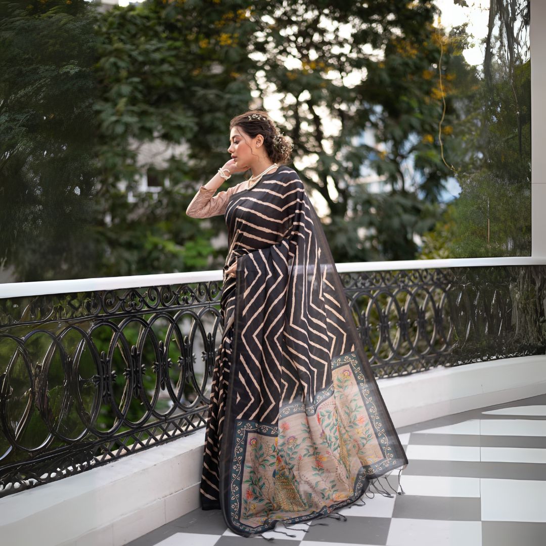 A woman models a striking Black Saree with a bold beige geometric print. The pallu is light beige with a dark border, featuring an elaborate printed motif of colorful flowers and foliage. She wears a gold necklace and is posed on an outdoor balcony.