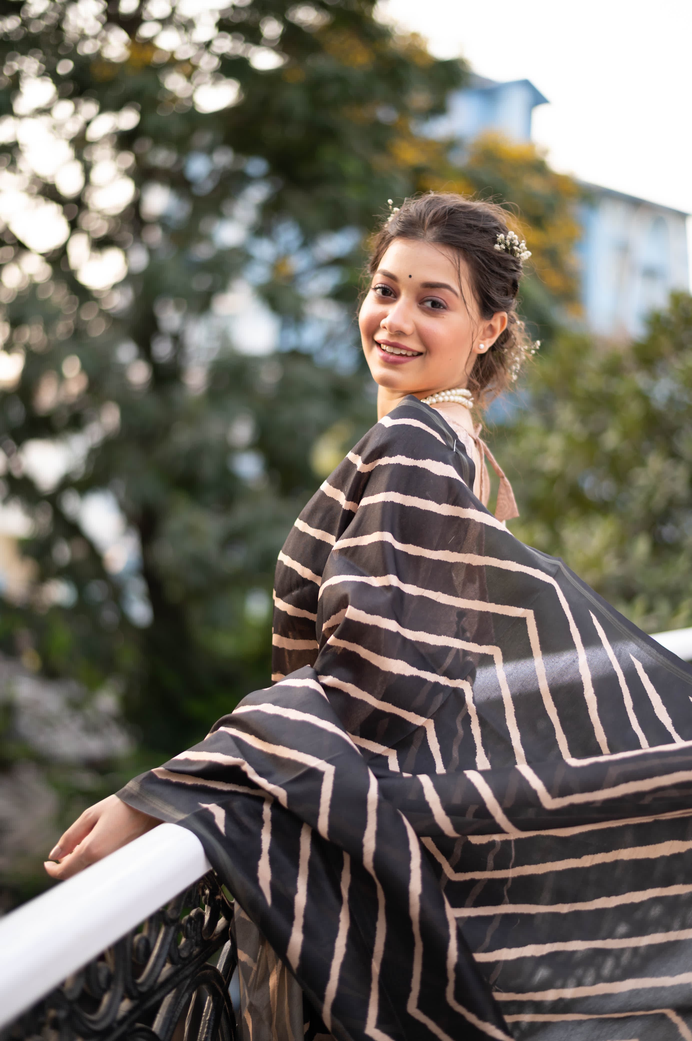 A woman models a striking Black Saree with a bold beige geometric print. The pallu is light beige with a dark border, featuring an elaborate printed motif of colorful flowers and foliage. She wears a gold necklace and is posed on an outdoor balcony.