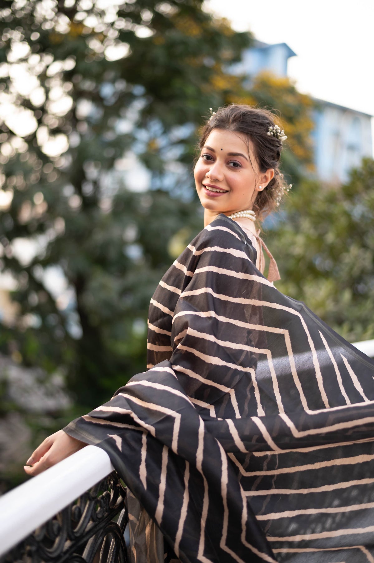 A woman models a striking Black Saree with a bold beige geometric print. The pallu is light beige with a dark border, featuring an elaborate printed motif of colorful flowers and foliage. She wears a gold necklace and is posed on an outdoor balcony.