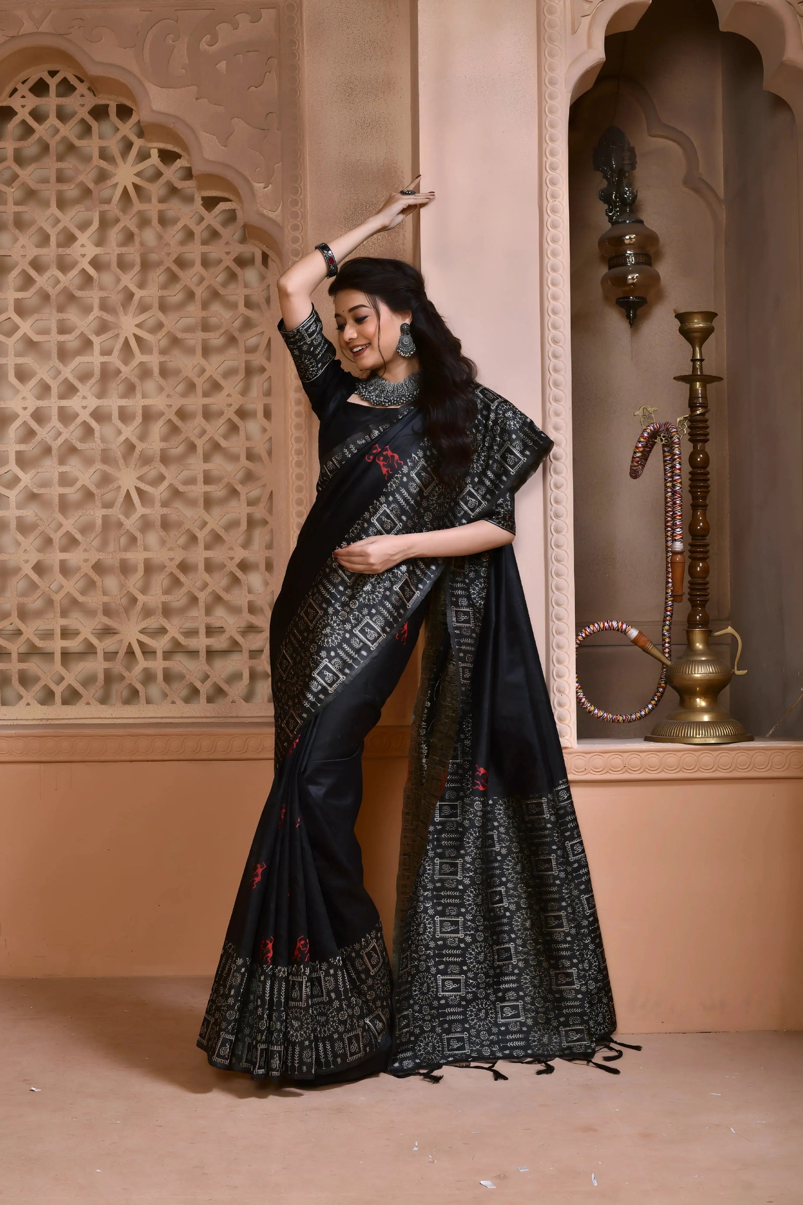 Woman wearing a black silk saree with intricate geometric border and subtle red motifs, paired with traditional silver jewelry, standing in a heritage-inspired room with ornate lattice window and carved walls
