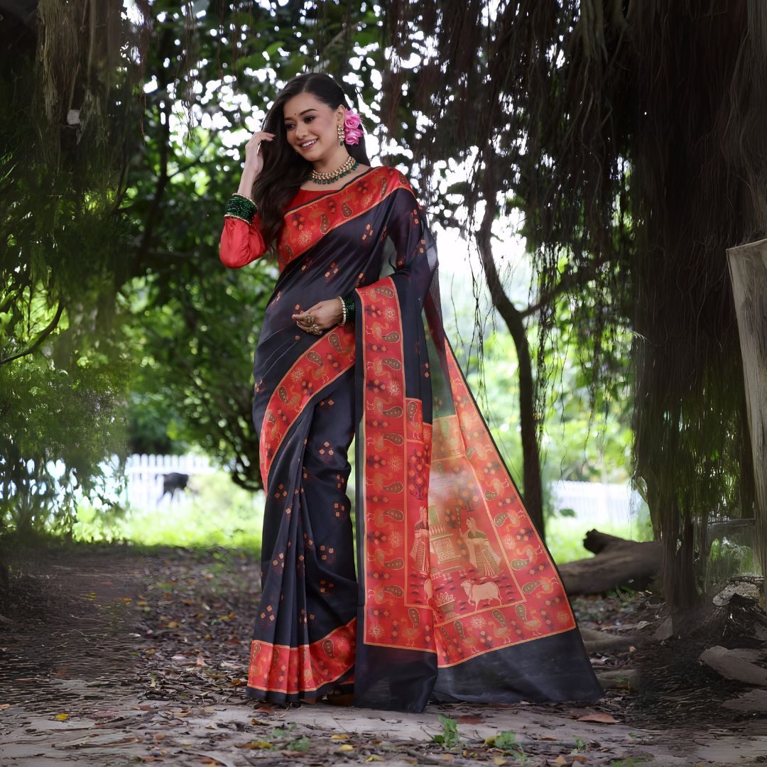 A woman models a striking Black Soft Tussar Silk Saree with small motifs. The saree has a wide, contrasting deep red border and pallu featuring intricate traditional and figurative prints. She wears a long-sleeve red blouse, a green beaded necklace, and is posed in a garden setting.