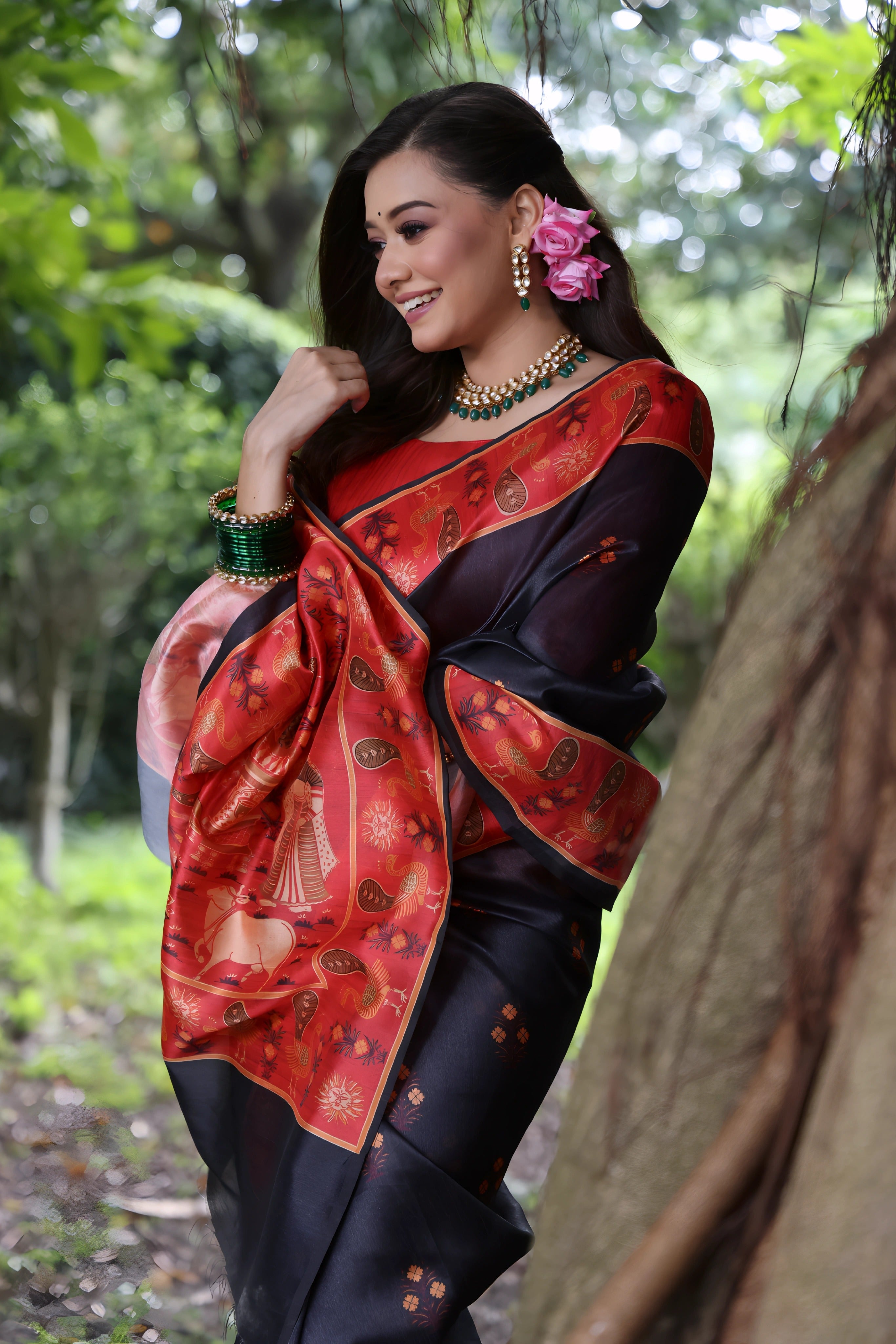 A woman models a striking Black Soft Tussar Silk Saree with small motifs. The saree has a wide, contrasting deep red border and pallu featuring intricate traditional and figurative prints. She wears a long-sleeve red blouse, a green beaded necklace, and is posed in a garden setting.