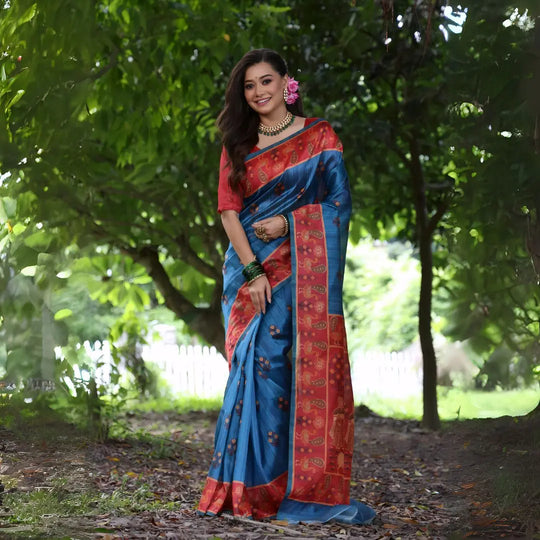 A woman models a vibrant Blue Soft Tussar Silk Saree with small motifs. The saree has a wide, contrasting deep red border and pallu featuring intricate traditional prints. She wears a red blouse, a green beaded necklace, and has a pink flower in her hair.