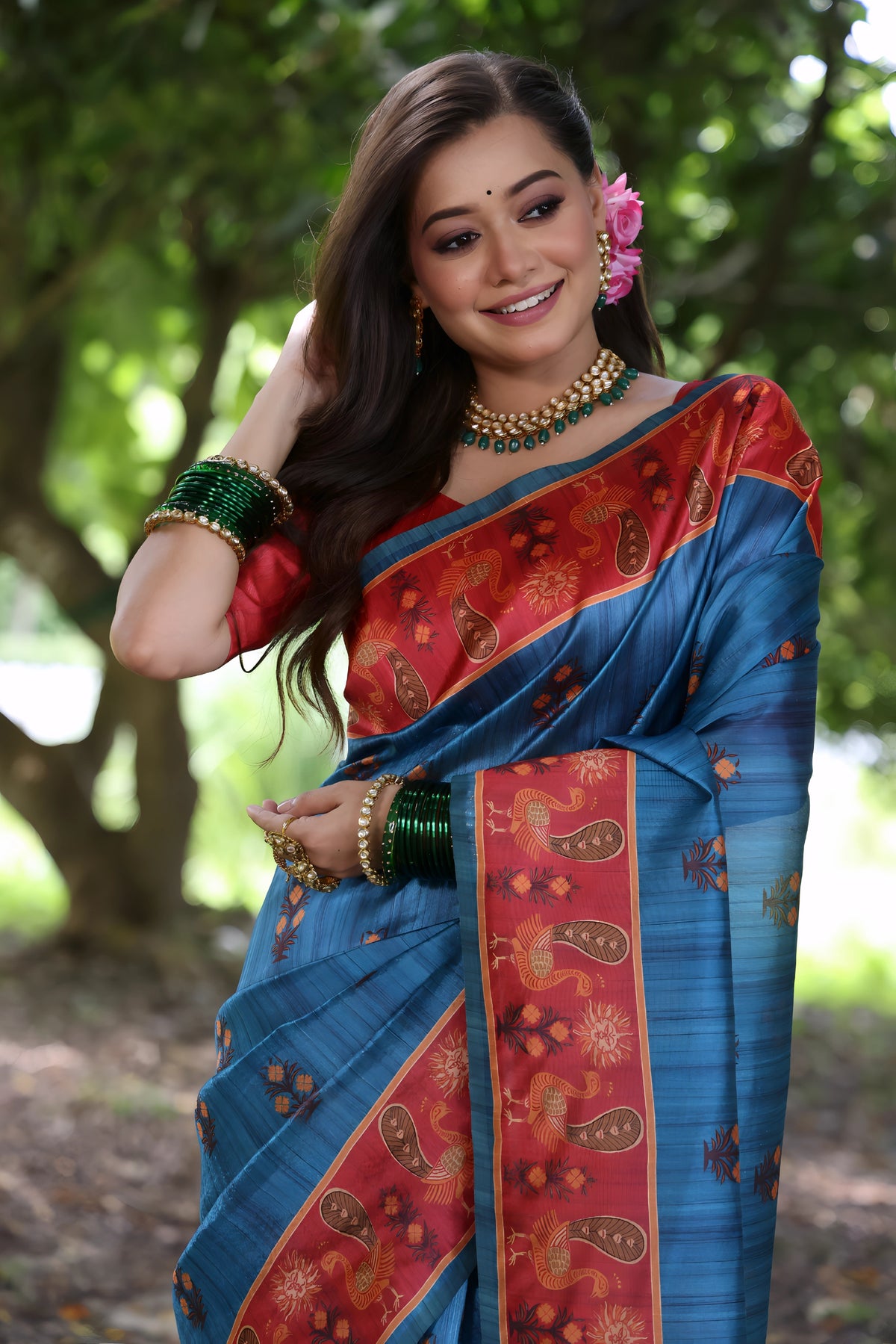 A woman models a vibrant Blue Soft Tussar Silk Saree with small motifs. The saree has a wide, contrasting deep red border and pallu featuring intricate traditional prints. She wears a red blouse, a green beaded necklace, and has a pink flower in her hair.