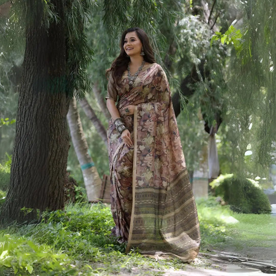 A woman models a Soft Tussar Silk Saree in a muted brown, covered in an all-over artistic print featuring floral motifs. The saree has a wide, woven Zari border with geometric patterns. She wears a complementary blouse and a substantial set of oxidized silver jewelry, posing against a large tree.