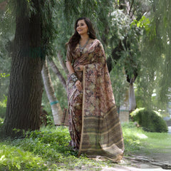 A woman models a Soft Tussar Silk Saree in a muted brown, covered in an all-over artistic print featuring floral motifs. The saree has a wide, woven Zari border with geometric patterns. She wears a complementary blouse and a substantial set of oxidized silver jewelry, posing against a large tree.