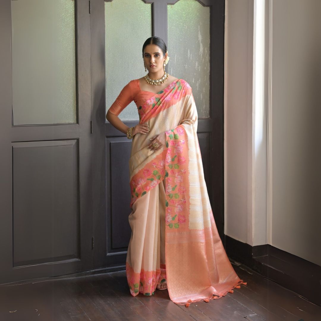 A woman models a graceful Soft Silk Saree. The saree body is pale beige/cream with subtle Zari weaving, and features a peach/coral border. The shoulder and pallu area are decorated with delicate printed floral motifs in pink and green. She pairs it with a peach-colored blouse and a gold and pearl choker necklace.