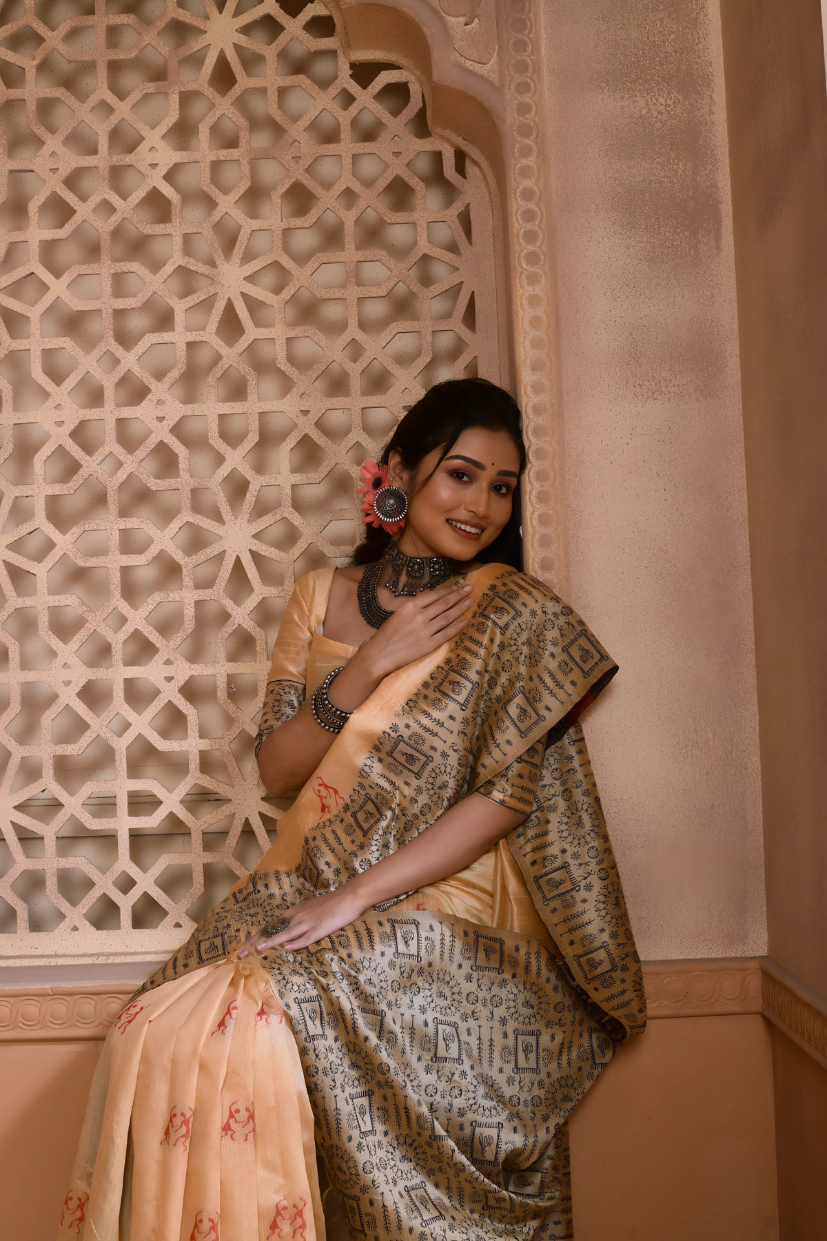 A woman models an elegant Beige Banglori Raw Silk Saree. The body features small motifs, and the pallu and border have dense, traditional Kalamkari-style weaving in a contrasting dark grey/black color. She wears a beige blouse and heavy oxidized silver jewelry, posing in an ornate, traditional setting.