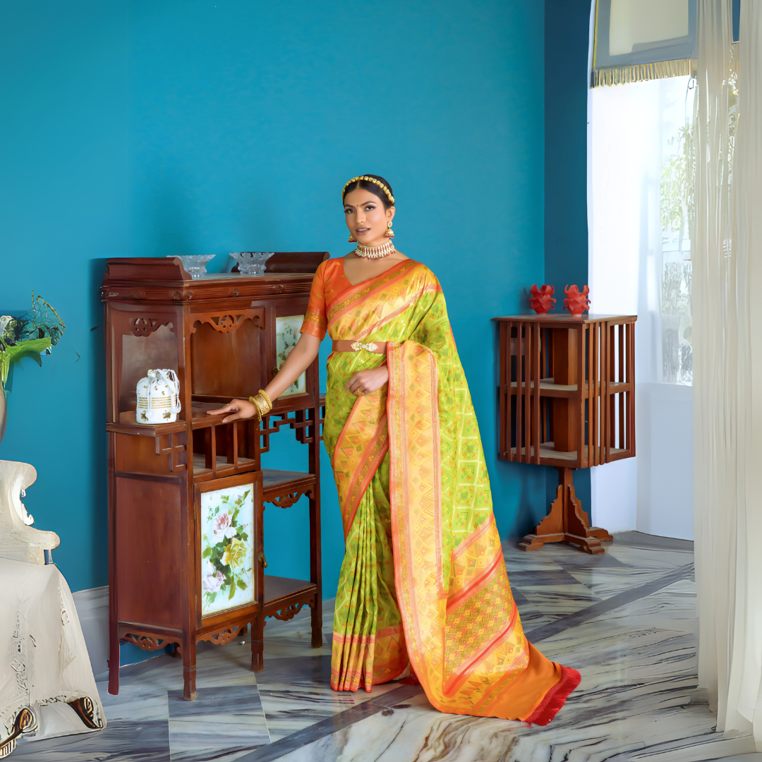 A woman models a striking Lime Green Banarasi Silk Saree with all-over Zari weaving. The wide pallu and border contrast dramatically in Bright Orange with large traditional Zari weaving patterns. She wears an orange blouse, traditional gold jewelry, and a metallic waist belt, posing indoors.