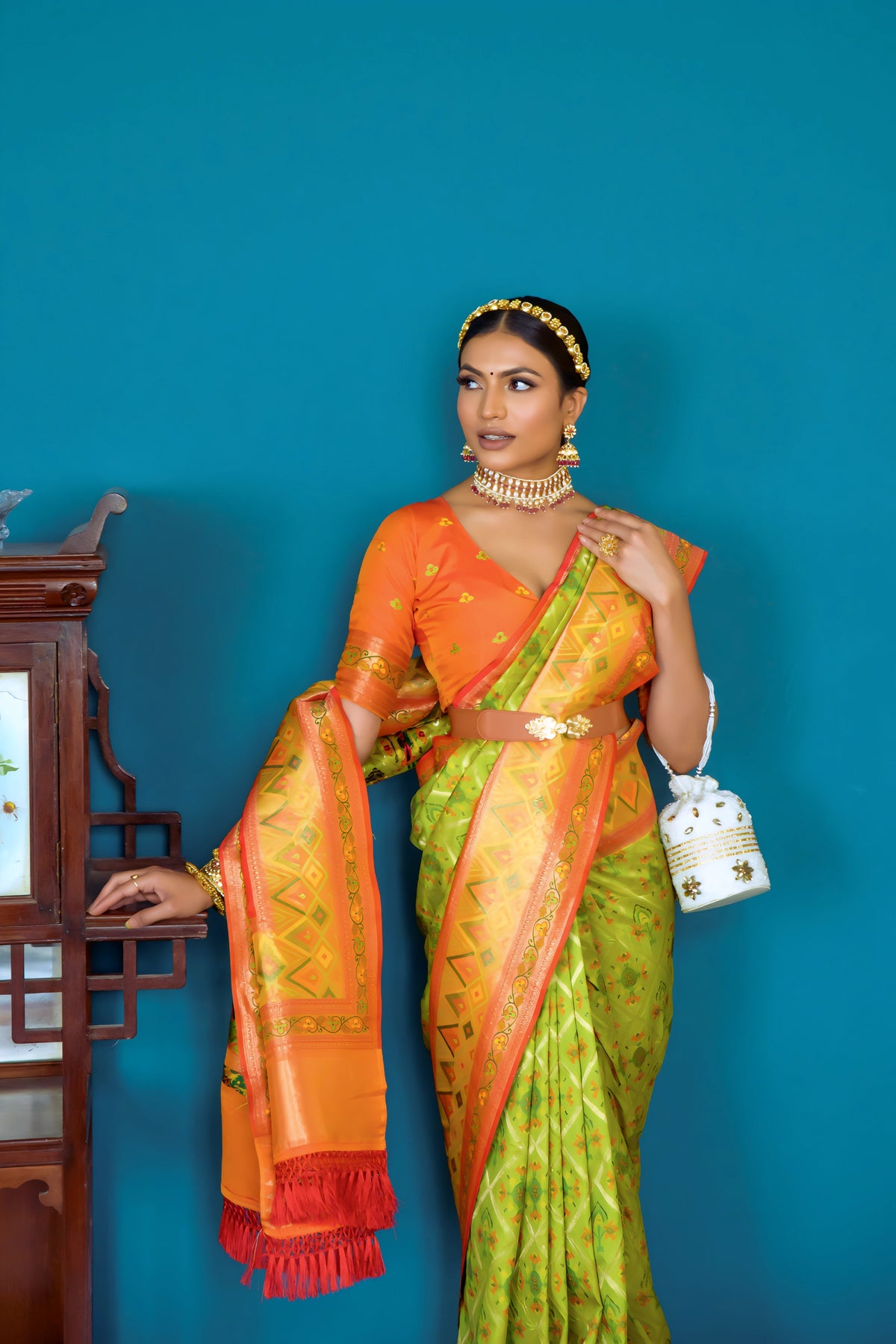 A woman models a striking Lime Green Banarasi Silk Saree with all-over Zari weaving. The wide pallu and border contrast dramatically in Bright Orange with large traditional Zari weaving patterns. She wears an orange blouse, traditional gold jewelry, and a metallic waist belt, posing indoors.