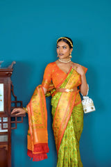 A woman models a striking Lime Green Banarasi Silk Saree with all-over Zari weaving. The wide pallu and border contrast dramatically in Bright Orange with large traditional Zari weaving patterns. She wears an orange blouse, traditional gold jewelry, and a metallic waist belt, posing indoors.