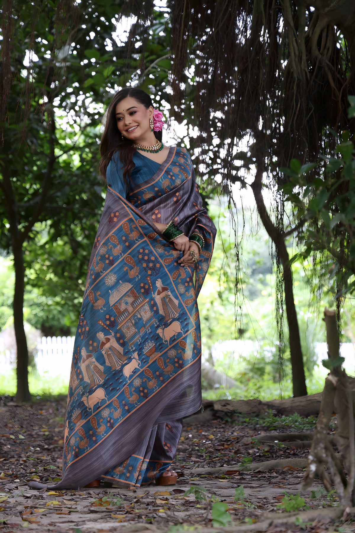 A woman models a sophisticated Soft Tussar Silk Saree. The saree is charcoal gray with a wide, contrasting deep teal and rust border and pallu featuring intricate traditional and figurative prints. She wears a teal blouse, a green beaded necklace, and is posed in a garden setting.