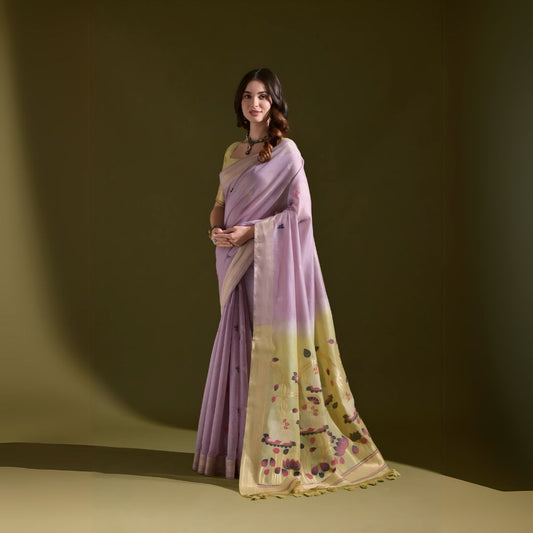 A woman models a Soft Muga Cotton Saree. The saree's body is light lavender, framed by a narrow Zari border. The pallu transitions into a pale yellow panel featuring colorful woven motifs. She pairs it with a light yellow blouse and a dark choker necklace.