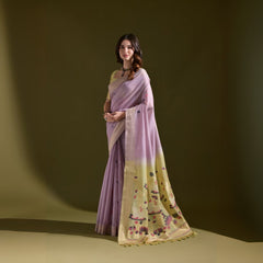 A woman models a Soft Muga Cotton Saree. The saree's body is light lavender, framed by a narrow Zari border. The pallu transitions into a pale yellow panel featuring colorful woven motifs. She pairs it with a light yellow blouse and a dark choker necklace.