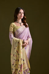 A woman models a Soft Muga Cotton Saree. The saree's body is light lavender, framed by a narrow Zari border. The pallu transitions into a pale yellow panel featuring colorful woven motifs. She pairs it with a light yellow blouse and a dark choker necklace.