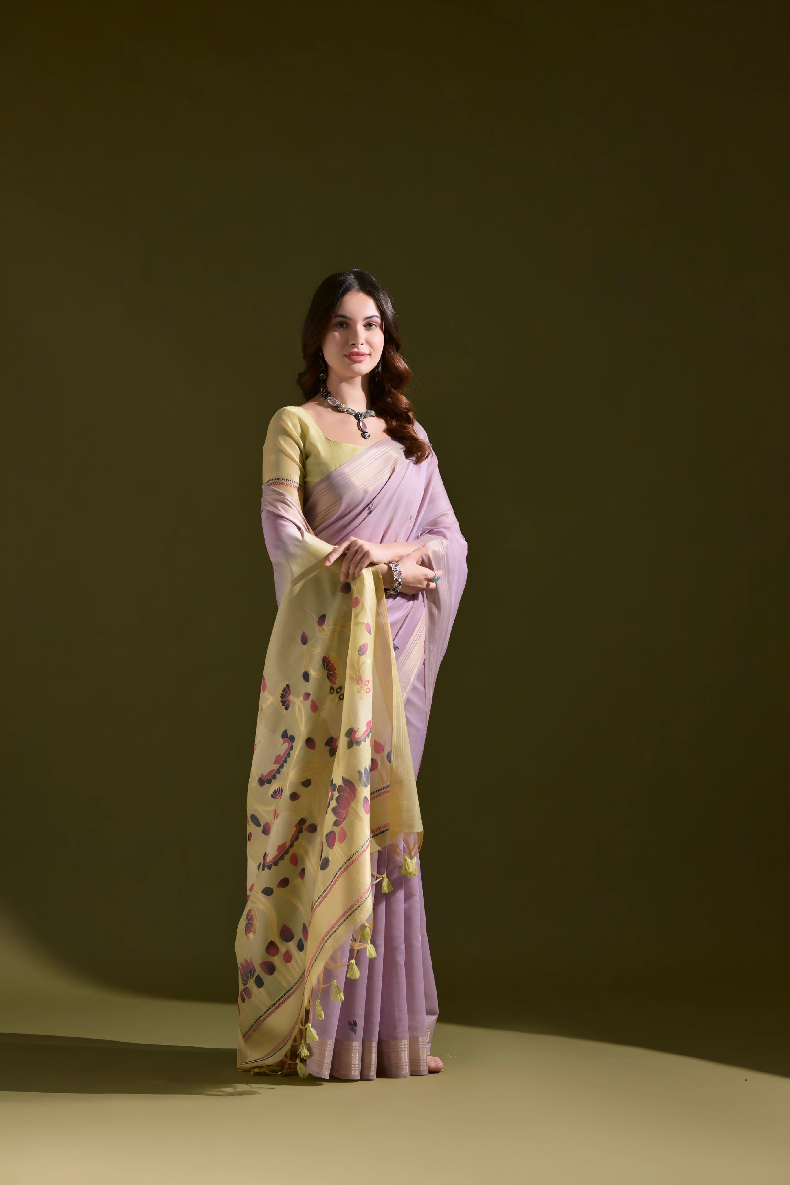A woman models a Soft Muga Cotton Saree. The saree's body is light lavender, framed by a narrow Zari border. The pallu transitions into a pale yellow panel featuring colorful woven motifs. She pairs it with a light yellow blouse and a dark choker necklace.